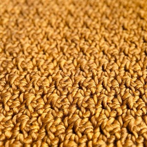 May include: Close-up of a textured, golden-brown crochet fabric. The detailed pattern shows a series of raised stitches, creating a dense, tactile surface. The warm color and intricate design suggest a handmade item, possibly a blanket or scarf.