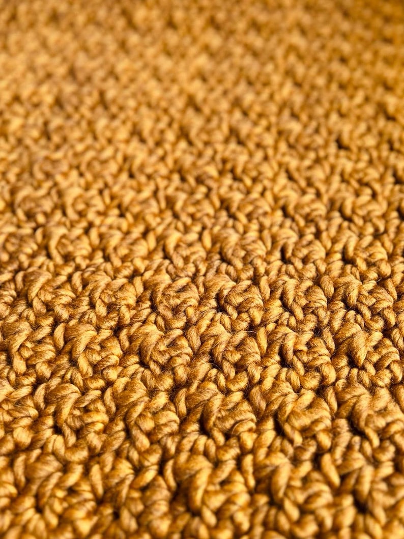 May include: Close-up of a textured, golden-brown crochet fabric. The detailed pattern shows a series of raised stitches, creating a dense, tactile surface. The warm color and intricate design suggest a handmade item, possibly a blanket or scarf.
