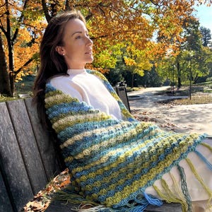 May include: A crocheted shawl featuring a textured, wavy pattern in alternating stripes of light blue, sage green, and cream. The rectangular wrap is finished with long, loose yarn fringe along the bottom edge in matching blue and green tones.