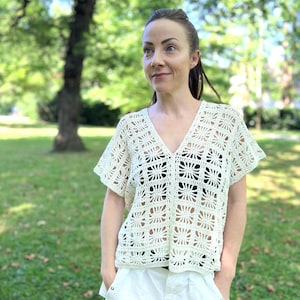 May include: Cream-colored, open-weave crochet top with a V-neck and short sleeves. The top has a floral pattern and is paired with white shorts. The background is a park with green grass and trees.