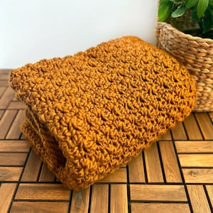 May include: Folded mustard yellow crochet blanket. The blanket is made of thick yarn and has a textured, woven appearance. The blanket is folded neatly and rests on a wooden surface. A wicker basket with a green plant is in the background.