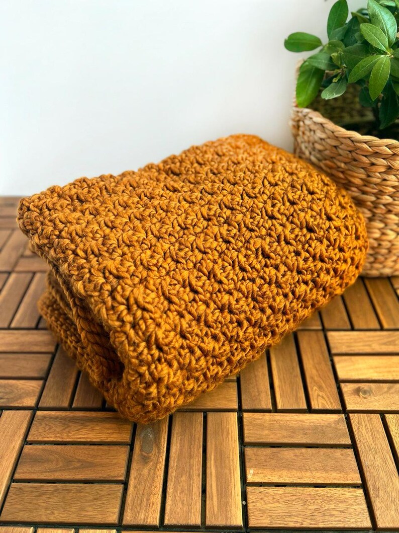 May include: Folded mustard yellow crochet blanket. The blanket is made of thick yarn and has a textured, woven appearance. The blanket is folded neatly and rests on a wooden surface. A wicker basket with a green plant is in the background.