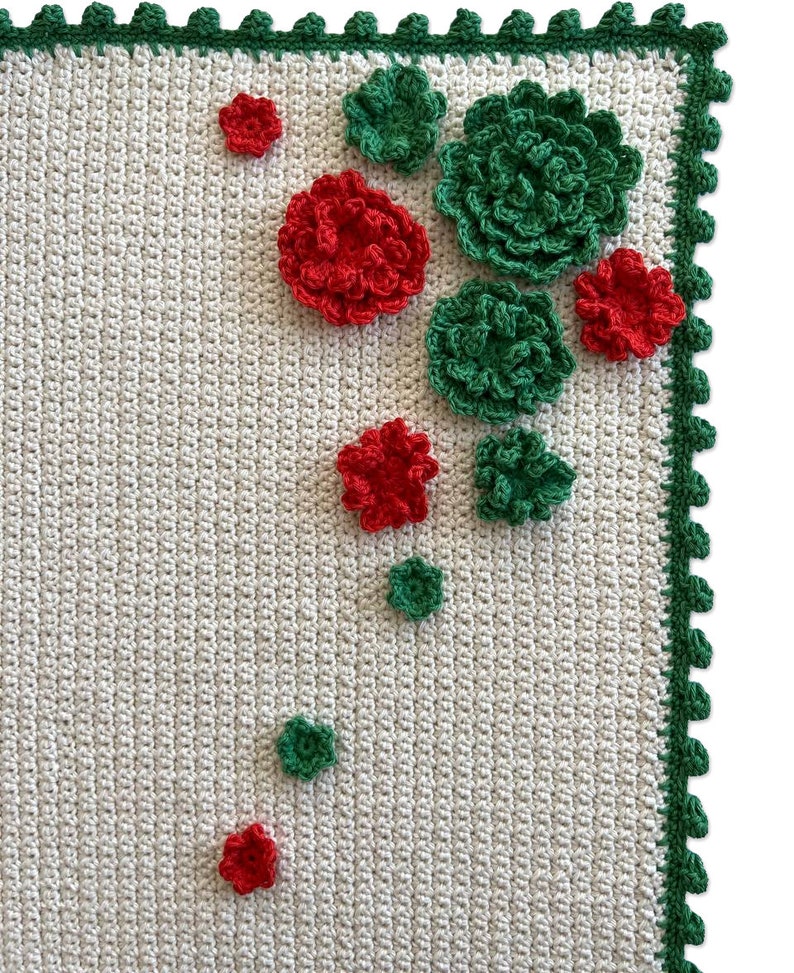 May include: A white crocheted blanket with a green border and red and green crocheted flowers.