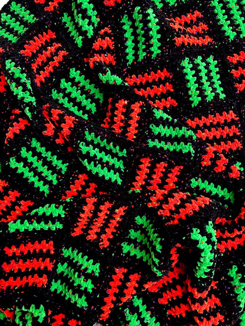 May include: Close-up of a crocheted textile with a repeating pattern. The design features vibrant neon green and red zigzags set against a black background. The texture appears soft and handmade, with a slightly irregular stitch.