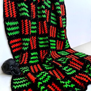 May include: A handmade crochet blanket in black, red, and green. The blanket features a repeating pattern of rectangular blocks with vertical stripes. A black skull figurine rests on the blanket. The blanket is made of soft yarn.