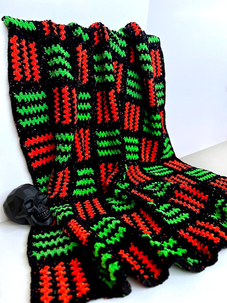 May include: A handmade crochet blanket in black, red, and green. The blanket features a repeating pattern of rectangular blocks with vertical stripes. A black skull figurine rests on the blanket. The blanket is made of soft yarn.