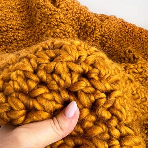 May include: Close-up of a mustard yellow, chunky knit blanket. The thick yarn is tightly woven, creating a textured surface. The blanket is folded, showing the depth and volume of the knitted material. The color is a warm, inviting shade.