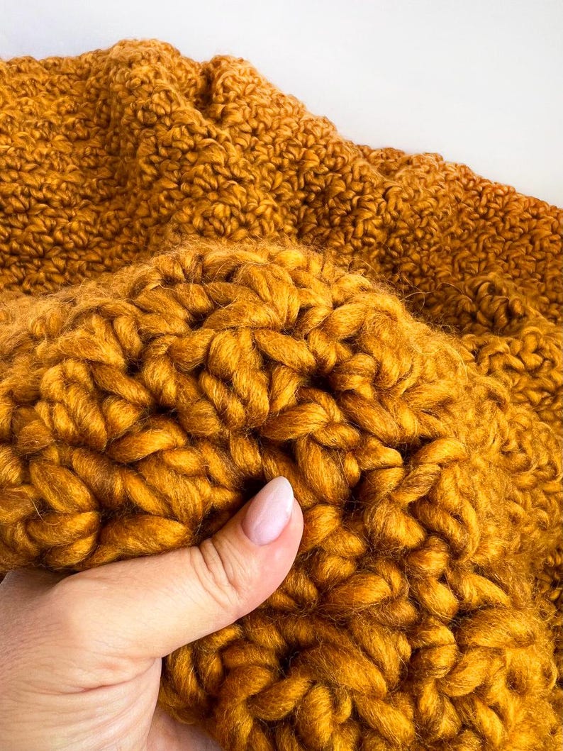 May include: Close-up of a mustard yellow, chunky knit blanket. The thick yarn is tightly woven, creating a textured surface. The blanket is folded, showing the depth and volume of the knitted material. The color is a warm, inviting shade.