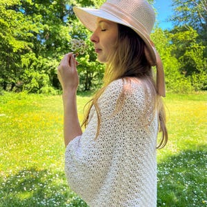 Może przedstawiać: Kobieta w kremowym szalu z otwartym splotem i kapeluszu ze słomy z szerokim rondem i różową wstążką. Kapelusz zacienia jej twarz, gdy trzyma mały bukiet białych kwiatów. Tło to słoneczna łąka z zielonymi drzewami.