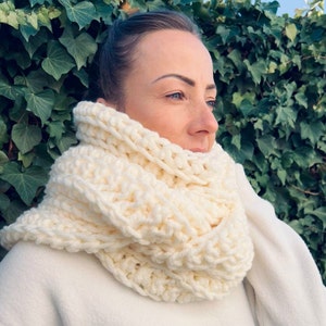 May include: A white crocheted infinity scarf. The scarf is thick and chunky, and it is worn by a person standing in front of a green ivy wall.
