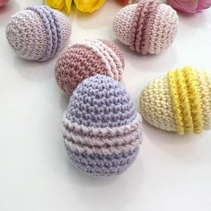 May include: Five crocheted Easter eggs in various pastel colors, including purple, pink, and yellow. The eggs are decorated with white crocheted stripes.