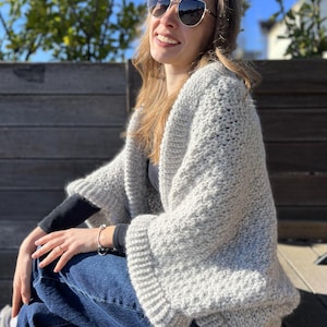 May include: A light gray, open-front crochet cardigan with wide sleeves. The cardigan is made of a chunky knit and has a ribbed hem and sleeve cuffs. The person is wearing blue jeans and sunglasses.