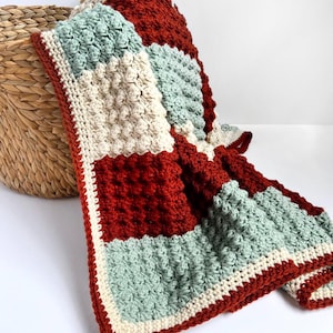 Pode incluir: Um cobertor de crochê feito à mão com um design de patchwork. O cobertor apresenta pontos texturizados em tons de vermelho, creme e verde-água. Uma borda vermelha escura emoldura o cobertor, que é dobrado sobre uma cesta de vime.