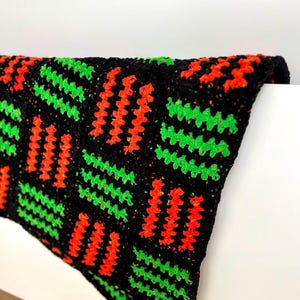 May include: A black crochet blanket with a repeating pattern of orange and green zigzags. The blanket is folded over a white surface, showcasing the textured design and vibrant colors. The blanket appears to be handmade.