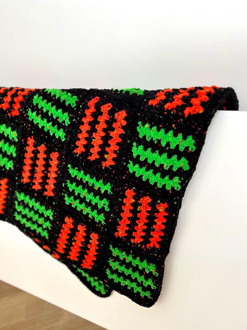 May include: A black crochet blanket with a repeating pattern of orange and green zigzags. The blanket is folded over a white surface, showcasing the textured design and vibrant colors. The blanket appears to be handmade.