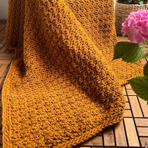 May include: Mustard yellow crocheted blanket with a textured stitch pattern. The blanket is folded over a chair and has a thick border. The blanket is made of a soft yarn and is perfect for snuggling up on the couch.