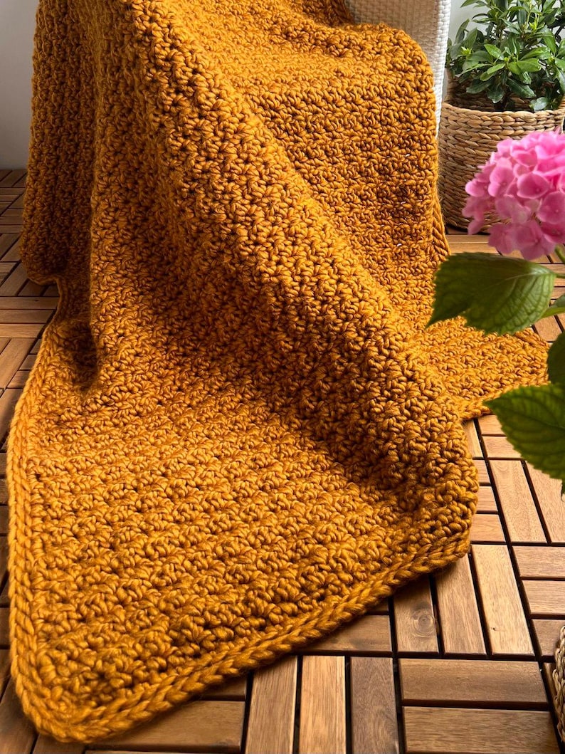 May include: Mustard yellow crocheted blanket with a textured stitch pattern. The blanket is folded over a chair and has a thick border. The blanket is made of a soft yarn and is perfect for snuggling up on the couch.