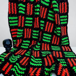 May include: A handmade crochet blanket in black, with a repeating pattern of vibrant red and green stitches. The blanket is folded, showcasing the texture and color contrast. A black skull figurine is visible in the background.