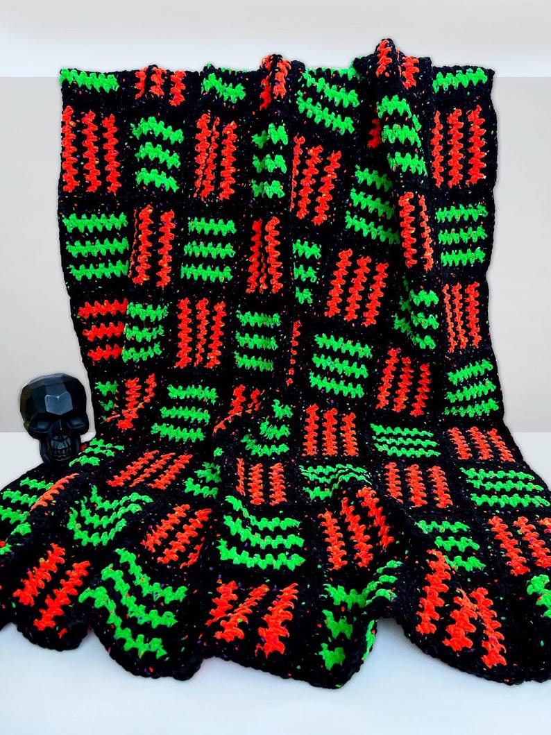 May include: A handmade crochet blanket in black, with a repeating pattern of vibrant red and green stitches. The blanket is folded, showcasing the texture and color contrast. A black skull figurine is visible in the background.