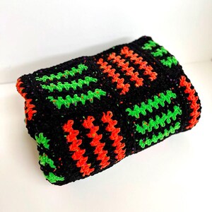 May include: A folded, rectangular crochet item in black, with vibrant neon green and orange geometric patterns. The item is likely a blanket or a decorative piece, showcasing a handmade aesthetic.