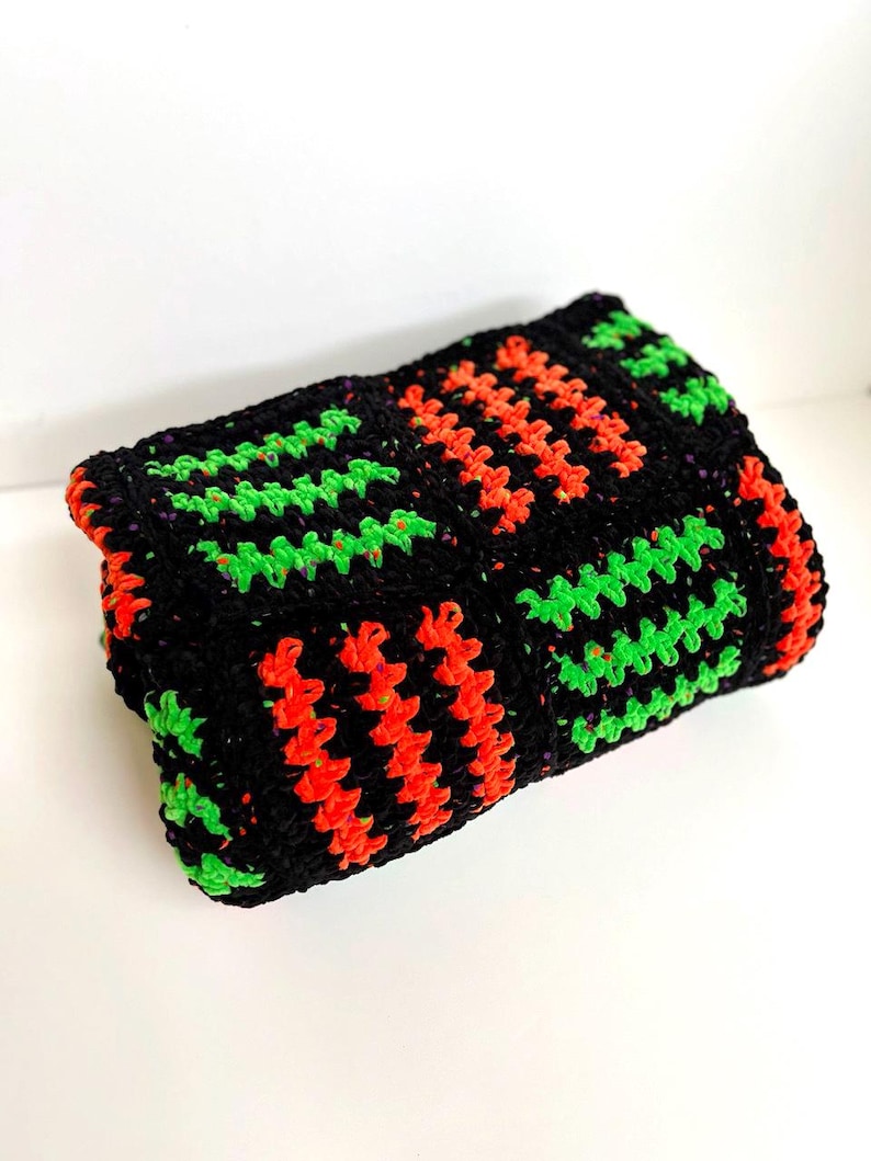 May include: A folded, rectangular crochet item in black, with vibrant neon green and orange geometric patterns. The item is likely a blanket or a decorative piece, showcasing a handmade aesthetic.