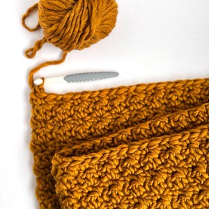 May include: Mustard yellow yarn ball and crochet project in progress. A white and gray crochet hook is placed near the project. The project is a thick, textured knit, likely a scarf or blanket, with a visible stitch pattern.