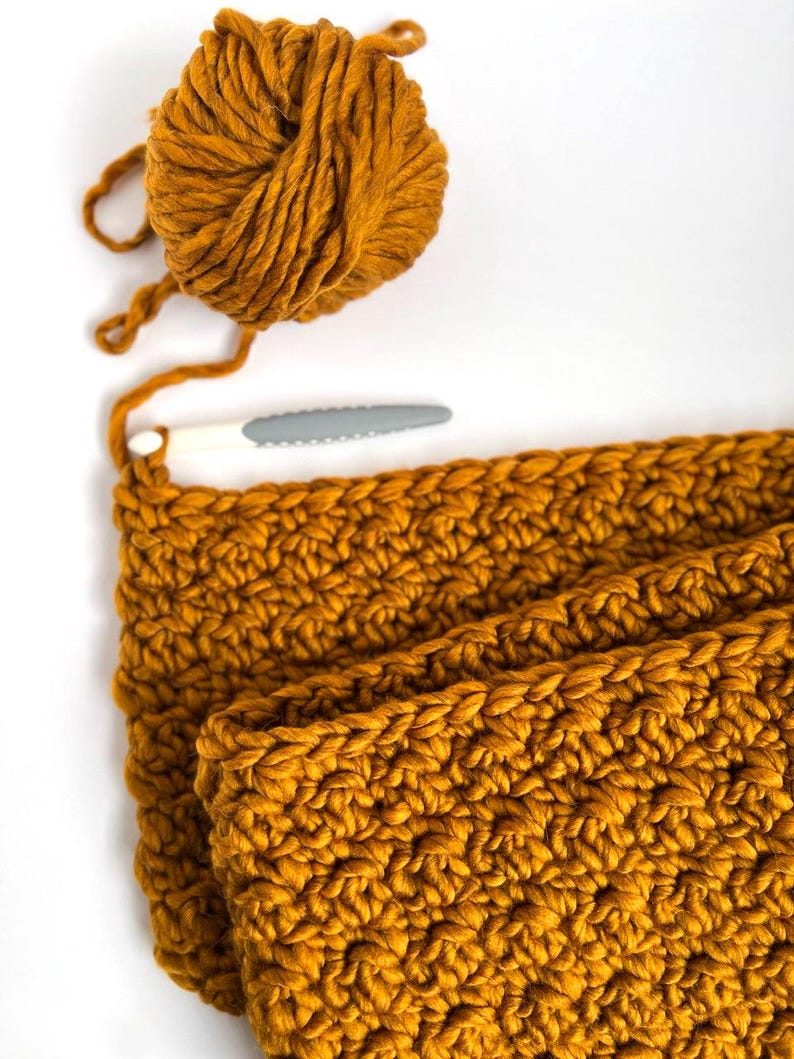 May include: Mustard yellow yarn ball and crochet project in progress. A white and gray crochet hook is placed near the project. The project is a thick, textured knit, likely a scarf or blanket, with a visible stitch pattern.