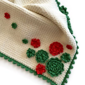 May include: A white crocheted baby blanket with a green crocheted border and red and green crocheted flowers.