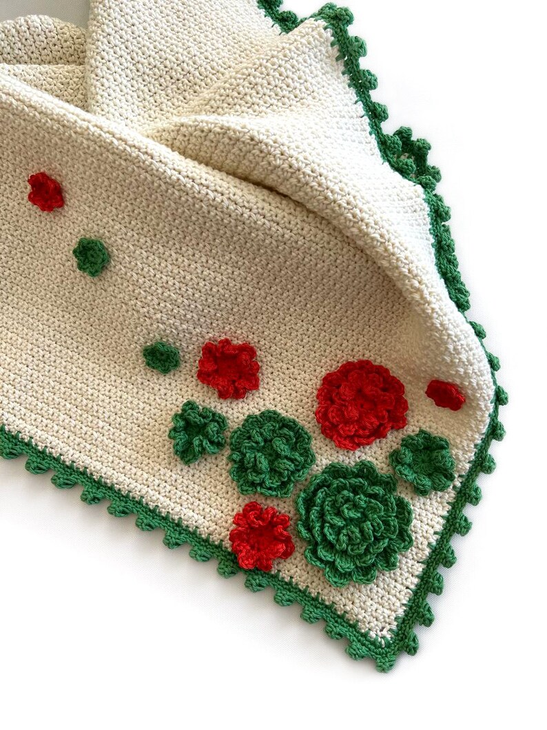 May include: A white crocheted baby blanket with a green crocheted border and red and green crocheted flowers.