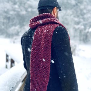 May include: A burgundy knit scarf with a textured pattern is draped over a person's shoulders. The person is wearing a dark jacket and a dark blue cap. Snowflakes are falling in a winter scene.