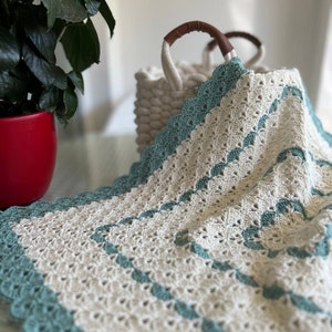 May include: A crocheted throw blanket with a white background and teal blue accents. The blanket has a scalloped edge and is folded over a wicker basket.