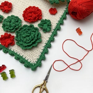 May include: A white crocheted blanket with green and red crocheted flowers, a ball of red yarn, a pair of gold scissors, and gummy bears.