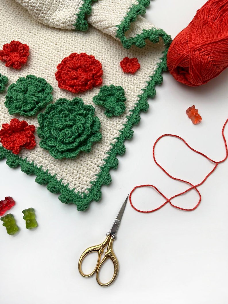 May include: A white crocheted blanket with green and red crocheted flowers, a ball of red yarn, a pair of gold scissors, and gummy bears.