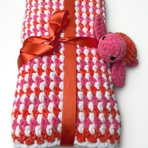 Peut inclure: Une couverture pour bébé en crochet rose et orange avec une bordure blanche, attachée avec un ruban rouge et un jouet en crochet de tortue rose.