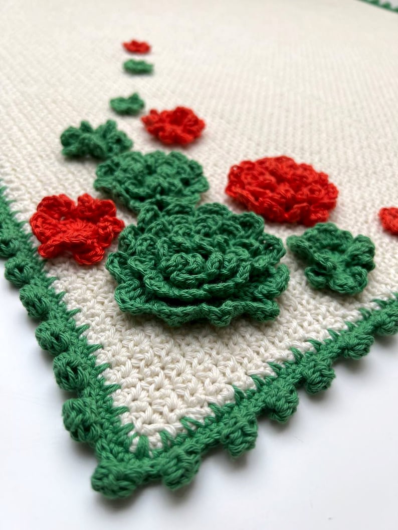 May include: A crocheted table runner with a white background and green and red crocheted flowers.