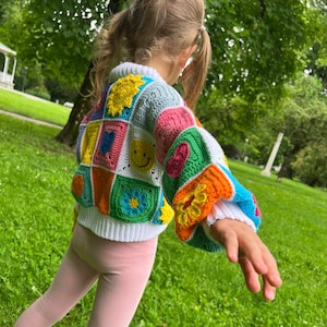 May include: A child wearing a colourful crocheted sweater with a patchwork design featuring flowers, smiley faces, and hearts. The sweater is white with a variety of bright colours, including red, yellow, blue, green, and orange.