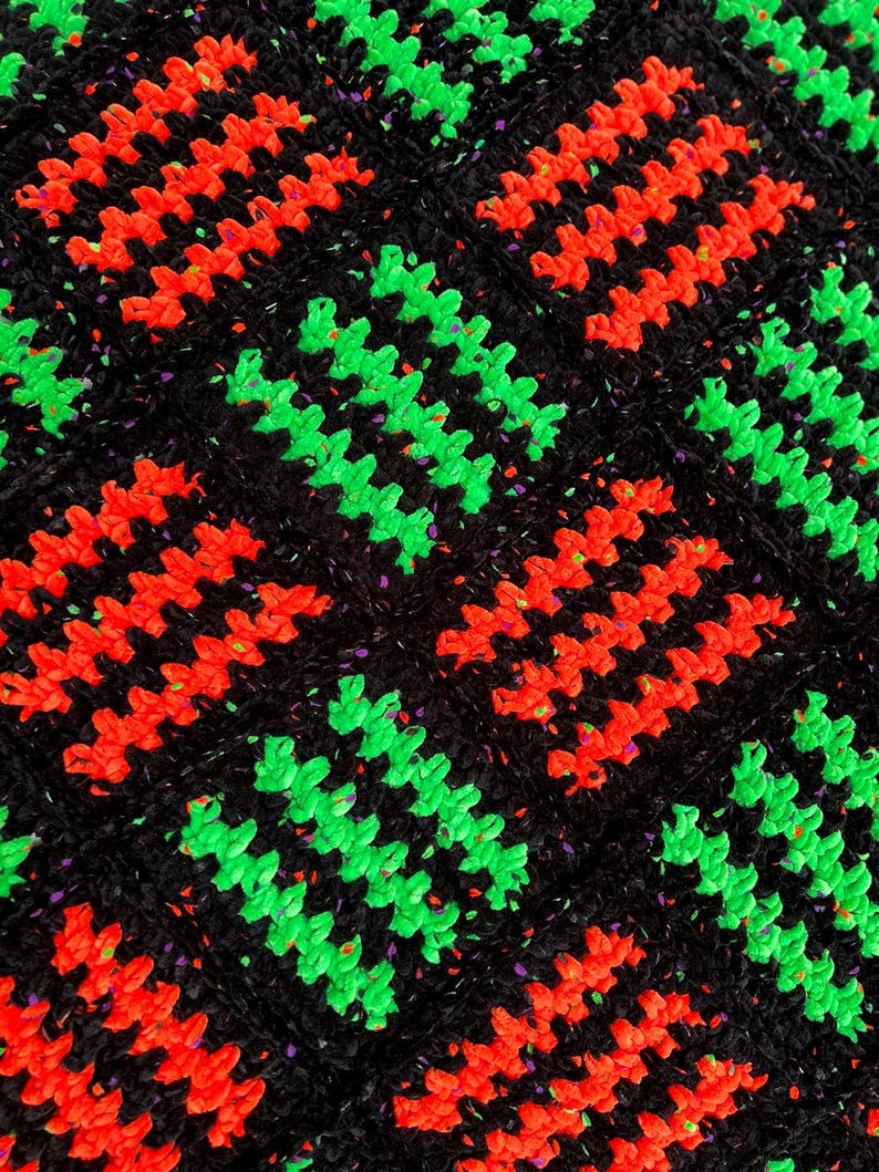 May include: Close-up of a crocheted textile with a repeating pattern. The design features rows of bright red and neon green zigzags set against a black background. The black yarn has flecks of other colors.