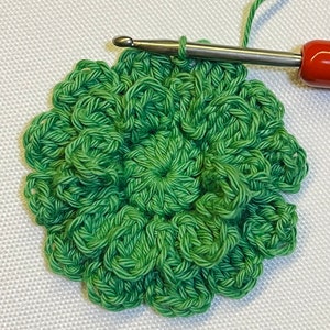 May include: A green crocheted flower with a center circle, being worked on with a crochet hook.