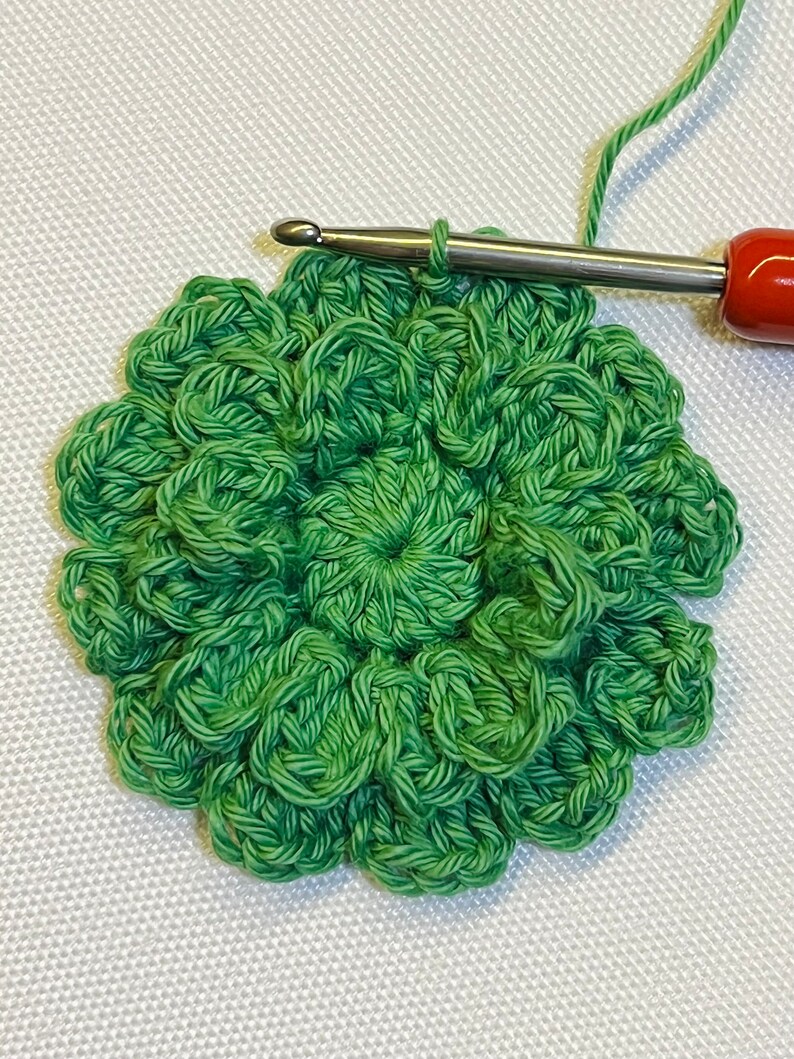 May include: A green crocheted flower with a center circle, being worked on with a crochet hook.