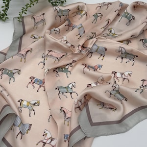 May include: A blush-colored silk scarf with a repeating pattern of illustrated horses in various colors and designs. The scarf has a gray border and is draped on a white surface.