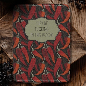 Peut inclure: Un carnet avec une couverture ornée d'un motif de piments rouges sur fond sombre. Le carnet porte l'inscription "THEY BE FUCKING IN THIS BOOK" dans un cadre vert clair. L'image est posée sur une surface en bois avec une bougie allumée.