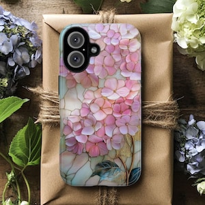以下が含まれることがあります： ピンクのアジサイの花柄とゴールドのアクセントが特徴の携帯電話ケース。茶色の紙で包まれたギフトの上に置かれ、新鮮な花と緑に囲まれ、自然で芸術的な美しさを演出しています。