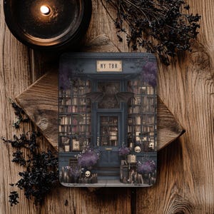 May include: A rectangular coaster featuring a dark blue bookstore facade with the text "MY TBR". The image includes a bat-wing design above the door, skulls, and purple floral accents. The coaster is set on a wooden surface.