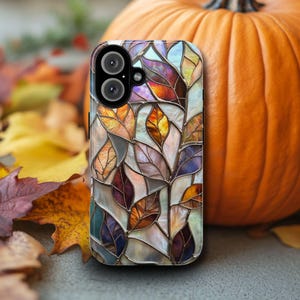 May include: A smartphone case with a stained glass leaf design in shades of orange, purple, and gold. The case is positioned in front of a large orange pumpkin and surrounded by fallen leaves, evoking an autumn theme.