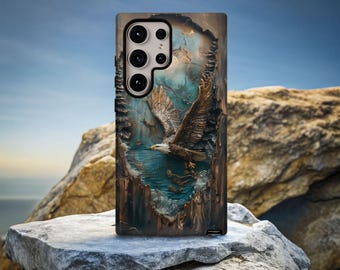 May include: A phone case featuring a detailed 3D relief of an eagle in flight over water. The case has a textured, wood-like appearance with shades of brown, blue, and white. The phone case is resting on a large rock with a blue sky background.