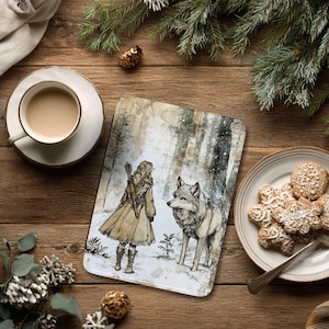 May include: A decorative tablet featuring a winter scene with a woman, a wolf, and a snowy forest. The artwork is in muted tones of beige, brown, and gray. The tablet is on a wooden surface, with a cup of coffee and cookies nearby.