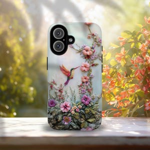 May include: A phone case featuring a 3D floral design with a hummingbird. The case has a light background with pink, purple, and white flowers. The hummingbird is depicted with red, orange, and purple feathers. The case has a dual-lens camera.