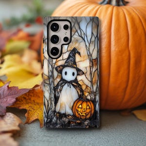 May include: A phone case featuring a Halloween-themed design. The case depicts a cartoon ghost wearing a witch hat and cape, holding a carved pumpkin. The background shows a forest scene with bare trees. An orange pumpkin is next to the phone case.