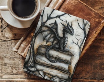May include: A fantasy-themed wallet featuring a black and grey dragon illustration on a stack of books. The background includes bare tree branches. A cup of coffee sits on a wooden surface nearby.
