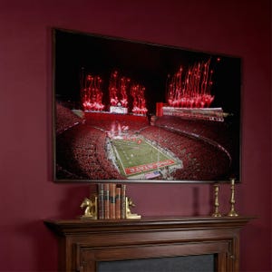 May include: A framed photograph of a football stadium at night, with a large crowd and red fireworks. The photo is displayed above a wooden mantelpiece. The mantelpiece has decorative items, including bookends and candlesticks.